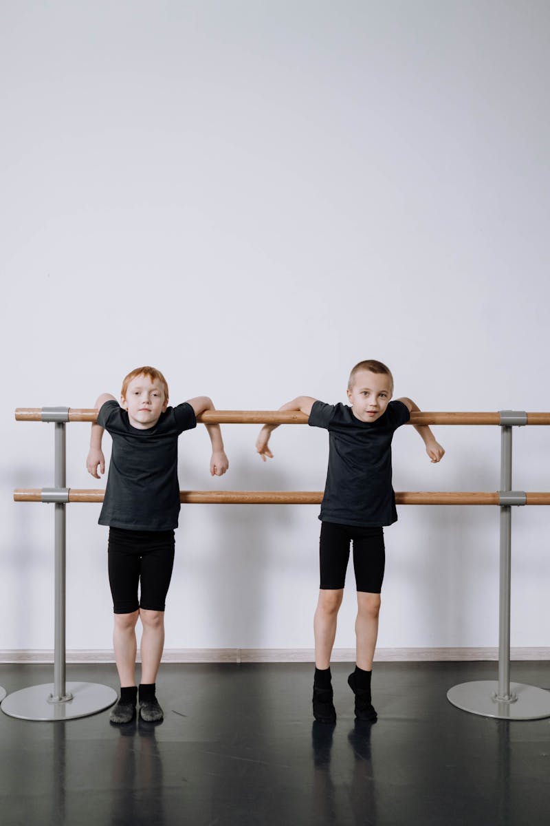 Two young boys stand at a ballet barre in a dance studio, wearing black attire, showcasing their interest in ballet.